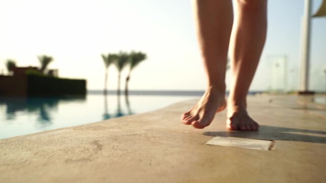 Feet step lightly near infinity pool edge. Warm sun glows over calm, endless water. Palm trees sway gently in background breeze. Feet move with care, hinting at relaxed vacation