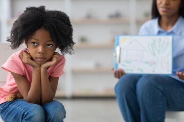 Fototapeta premium Worried Black Girl With Sad Expression Sitting On Floor While Listening To Therapist At Clinic. Engaged In A Session Focused On Emotional Support And Wellbeing, Weight Loss Concept.
