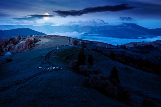 scenic carpathian autumn night. full moon over countryside mountain landscape with forest in fall colors. path leading through rolling hills in to the foggy valley under mysterious sky