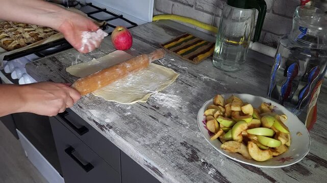 Woman rolling out pastry dough on a kitchen counter with a baked apple pie in the background.