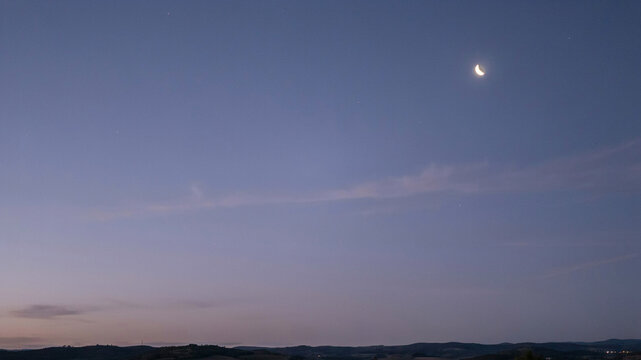 A serene twilight sky with a slender crescent moon and delicate, streaky clouds, showing a gradient from deep blue to soft lavender, above a dark, silhouetted rolling terrain.keywords: twilight