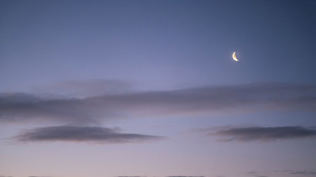 A thin, bright crescent moon is visible in a soft, gradient sky transitioning from pale blue to dusky purple, with delicate, horizontal streaks of grey clouds adding texture and depth to the serene