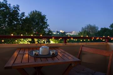 Naklejka premium Cozy balcony setup at twilight with warm string lights on the railing, a wooden table with ceramic teapot and cups, offering a serene city view for a relaxing evening atmosphere.