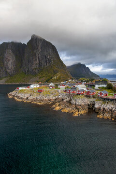 Malerische K&uuml;stenlandschaft von Hamn&oslash;y mit traditionellen roten Fischerh&uuml;tten, felsiger K&uuml;ste und dem imposanten Festh&aelig;ltinden unter dramatischem Wolkenhimmel, Hamn&oslash;y, Lofoten, Norwegen, Europa