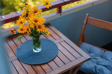 Naklejka premium Bouquet of bright yellow flowers in a glass vase on a wooden balcony table, featuring a cozy outdoor patio setting with warm sunlight and a wooden chair with cushion.
