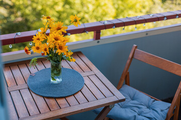 Naklejka premium Bright yellow flowers in a glass vase on a wooden balcony table with a cozy chair, placemat, string lights, and warm sunlight creating a charming outdoor setting. Autumn day