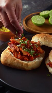 Hand sprinkling fresh herbs and chopped onion on Mexican al pastor pork sandwich with pineapple and lime