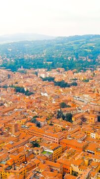 Vertical video. Bologna, Italy. the medieval Old Town, dense terracotta rooftops and historic street grid unfolding beneath a bright summer sky, with the green Apennine foothills rising beyond the ur