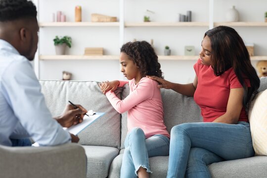 Worried Young Black Girl Sitting On Sofa With Mother In Therapy Session, Engaging In Conversation With Therapist In Office, Seeking Support And Guidance In A Counseling Environment.