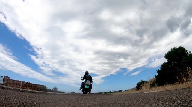 Slow motion fixed low angle view of a biker starting a custom motorcycle, entering a rural road and riding away into the distance.
