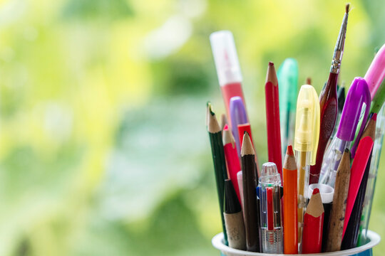 Colored pencils in a glass cup on a wooden table against a window with lush green foliage