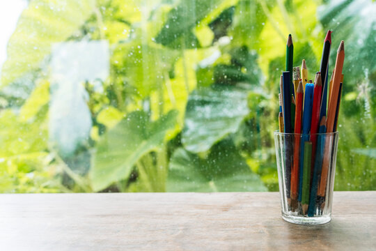 Colored pencils in a glass cup on a wooden table against a window with lush green foliage