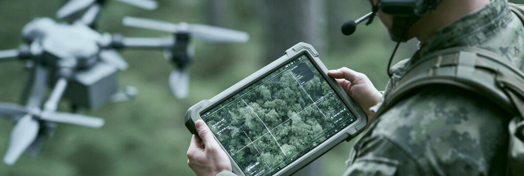 Male soldier operating a military drone with a tablet in a forest setting.