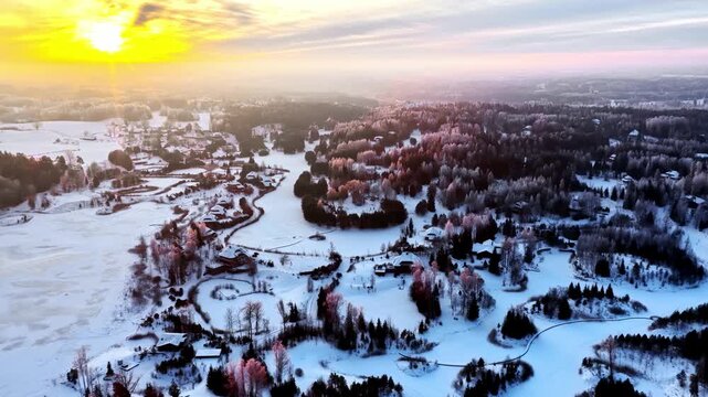 Aerial view of a snowy landscape with pine trees and houses during a golden sunset in Latvia.