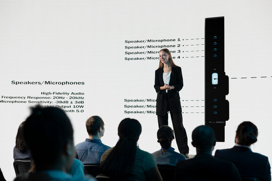 Young adult presents a mobile phone announcement on stage with promotional visuals, advertising a new release and investment deal during a business convention and product launch event for investors.