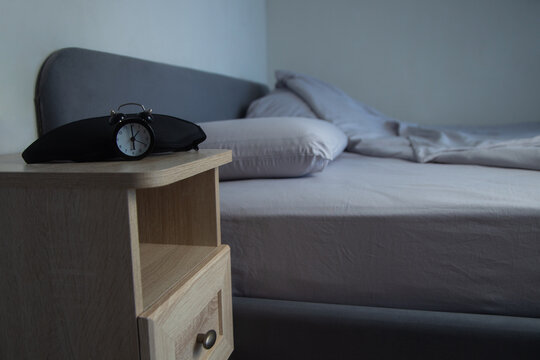 On a light wooden bedside table lies a black sleep mask with a miniature black alarm clock on the background of a bed with gray pastel linens. The concept of a sleep preparation atmosphere. High