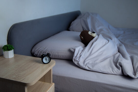 There is a mini alarm clock and a white potted plant on the bedside table, and a brown teddy bear is lying on a gray bed under a blanket in the background. The concept of a sleep mode, a comfortable