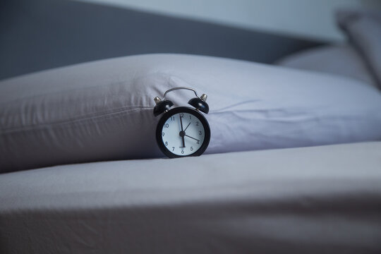 Close-up of a miniature black alarm clock showing 6 a.m. on a bed with gray pastel linens next to a pillow. Concept: morning time, early rise, cozy bedroom atmosphere. High quality photo