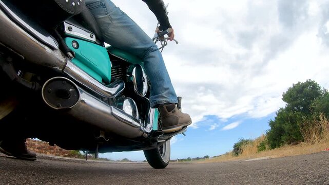 Slow motion low angle view of a biker slowing down and stopping a custom motorcycle, focusing on the lower part of the motorcycle and exhaust pipes.