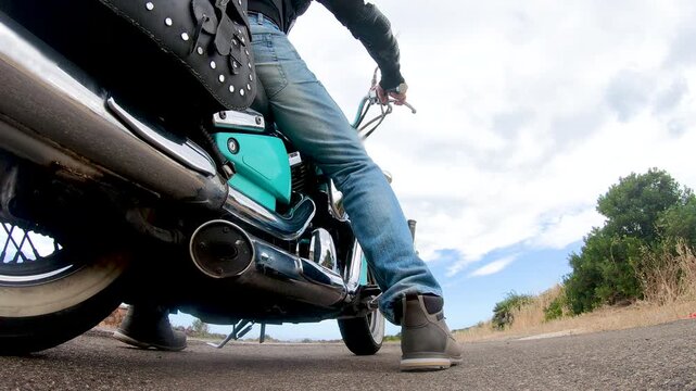 Slow motion low angle view of a biker mounting a turquoise custom motorcycle outdoors, focusing on the motorcycle and rider from the waist down in a cinematic lifestyle scene.