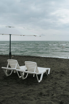 Empty white sun loungers and umbrella on a dark sand beach under a cloudy sky