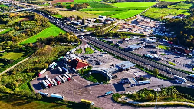 Aerial view of a highway rest stop featuring numerous parked trucks and service facilities surrounded by rural fields and greenery, Italy.