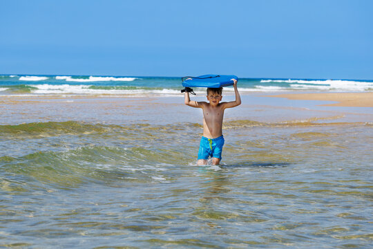 Child boy, carrying blue bodyboard through shallow ocean water