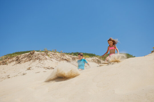 Boy and girl sliding down sandy dunes beneath clear blue sky