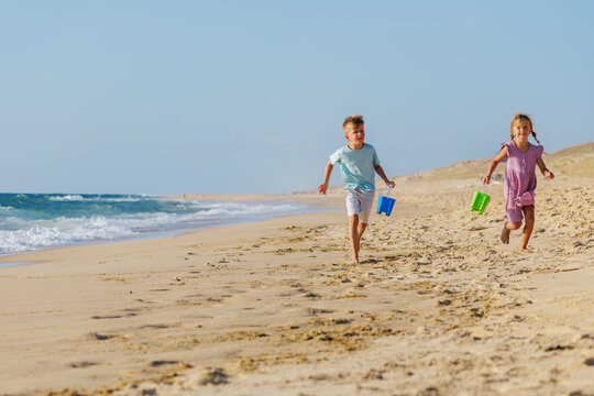 Happy kids running along sandy ocean beach with colorful buckets