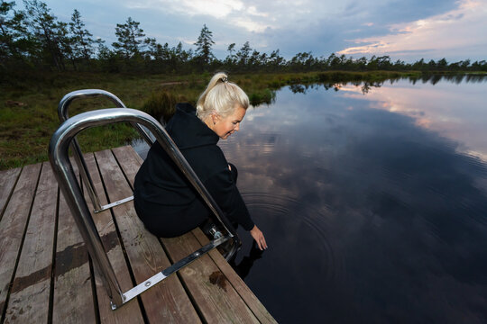 Blonde woman sitting on a wooden dock testing the cold water temperature of a tranquil bog lake in Estonia during late summer.