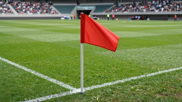 Red soccer corner flag stands at field edge with spectators in stadium background.