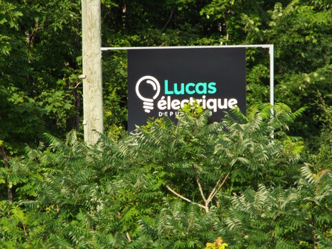 Gatineau, Quebec, Canada &ndash; June 30, 2026: A black sign reading "Lucas &eacute;lectrique" featuring a lightbulb logo is partially obscured by green leafy bushes outdoors on Rue de B&eacute;cancour.