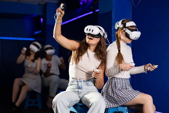 Cheerful young female friends wearing VR headsets and holding controllers having fun in virtual reality studio, competing in two-on-two shooter game