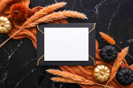 Halloween stationery mockup with a blank white card anf black envelope on a dark surface, decorated with pumpkins and dried pampas grass