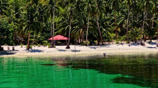 Aerial view of Port Barton Beach with lush palm trees, a red-roofed shelter on the white sand, and clear turquoise water in Mimaropa, Philippines.