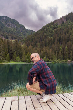 Senior man crouching on wooden deck by a lake, surrounded by mountains and forest