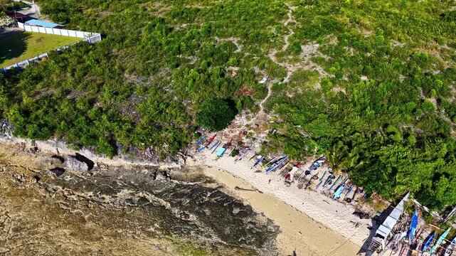Aerial view of traditional outrigger boats lined up on the sandy beach with lush green hillside and rocky coastline in Moalboal, Central Visayas, Philippines.