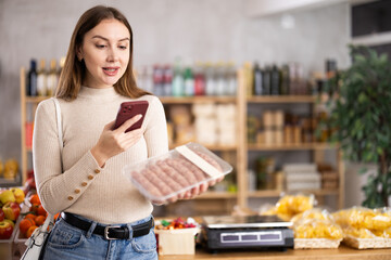 Young woman in store examines raw pork sausages in plastic container, scan QR-code. Buyer examines...