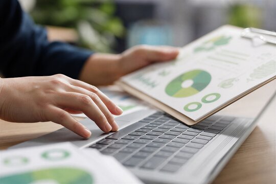 Close-up of hands typing on a laptop keyboard, holding an ESG report for corporate compliance and environmental sustainability data entry at an office desk.