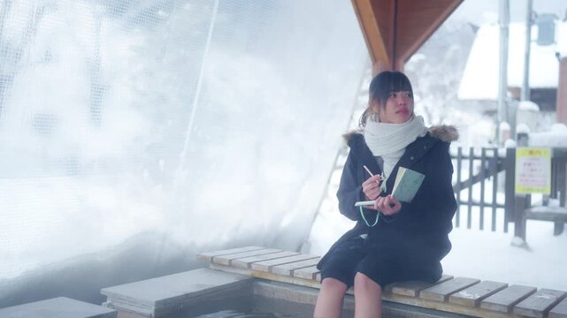 Asian woman writing travel diary while relaxing at outdoor hot spring foot bath in snowy winter. Female traveler enjoy outdoor lifestyle taking a break at highway rest area during road trip in Japan