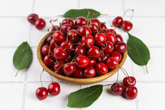 Top view of fresh ripe red sweet cherries in wooden bowl with green leaves on white tile textured background. Organic summer fruit, juicy berries, natural freshness, healthy eating, seasonal food
