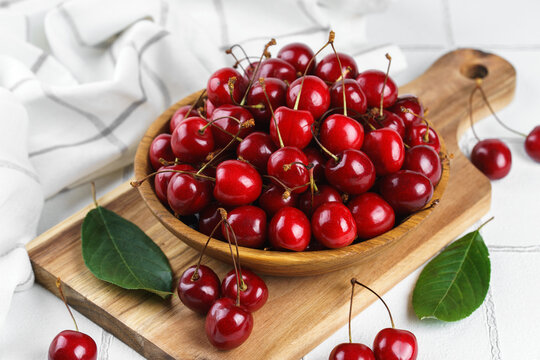 Fresh ripe sweet cherries in wooden bowl on rustic cutting board with green leaves and linen cloth on white textured background. Healthy summer fruit, red berries, fresh seasonal harvest, healthy food