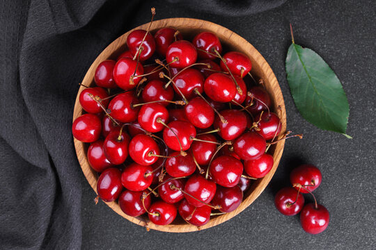 Fresh ripe red sweet cherries in wooden bowl on dark textured stone background with green leaves and black linen towel. Summer fruit, berry, harvest, healthy organic food