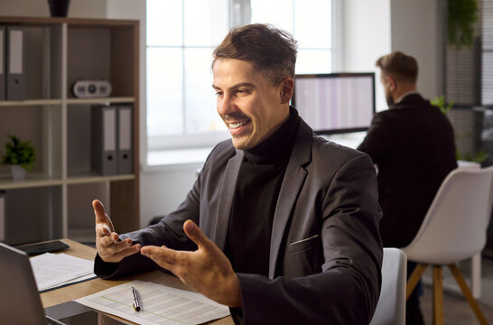 Business man on video call at office. He smiles and gestures to a client on a laptop discussing strategy, project updates and sales goals during an online meeting. Clear online team call idea.