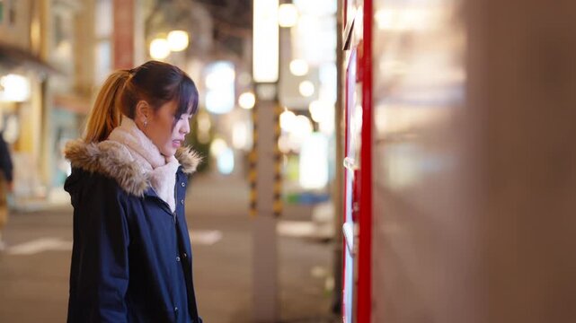 Young Asian woman choosing and buying drinks from beverage vending machine in Tokyo, Japan at night. Female tourist exploring city street, winter travel, sightseeing and holiday vacation.