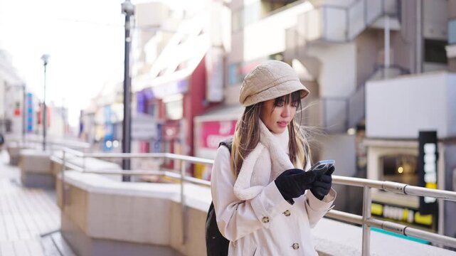 Young Asian woman traveler using smartphone searching navigation map while traveling in Tokyo city, Japan. Female tourist checking directions on mobile phone app during winter urban solo travel.