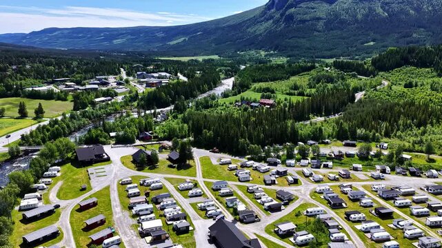 Aerial view of a large campground with numerous caravans and small cabins situated in a lush green valley surrounded by mountains and a river in Norway.