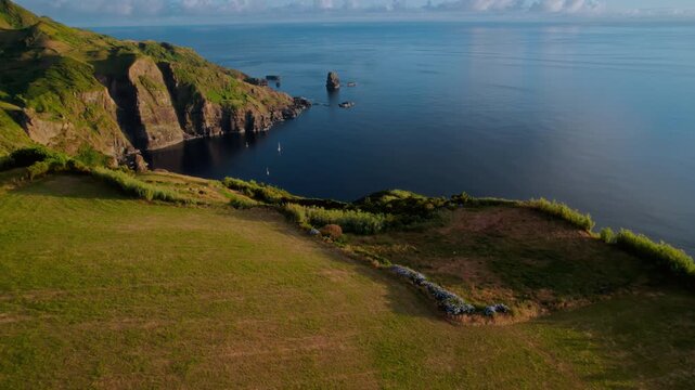 Beautiful high-angle aerial drone shot of a scenic coastal bay during morning golden hour, featuring lush green agricultural fields and rolling pastures on the edge of a high cliff looking down onto a