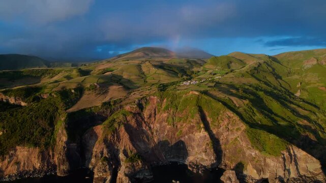 Wide aerial drone view of a dramatic volcanic coastline at golden hour, featuring steep rugged cliffs, a small rural village nestled in lush green rolling pastures divided by ancient stone walls, and