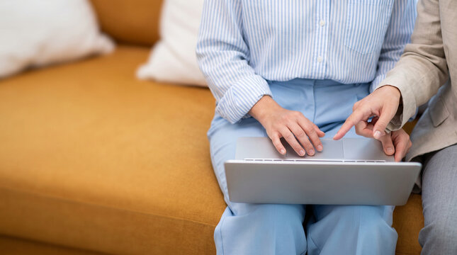 Cropped shot of two businesswomen working on laptop computer while sitting on sofa, colleague pointing at touchpad or screen, teamwork and digital collaboration concept